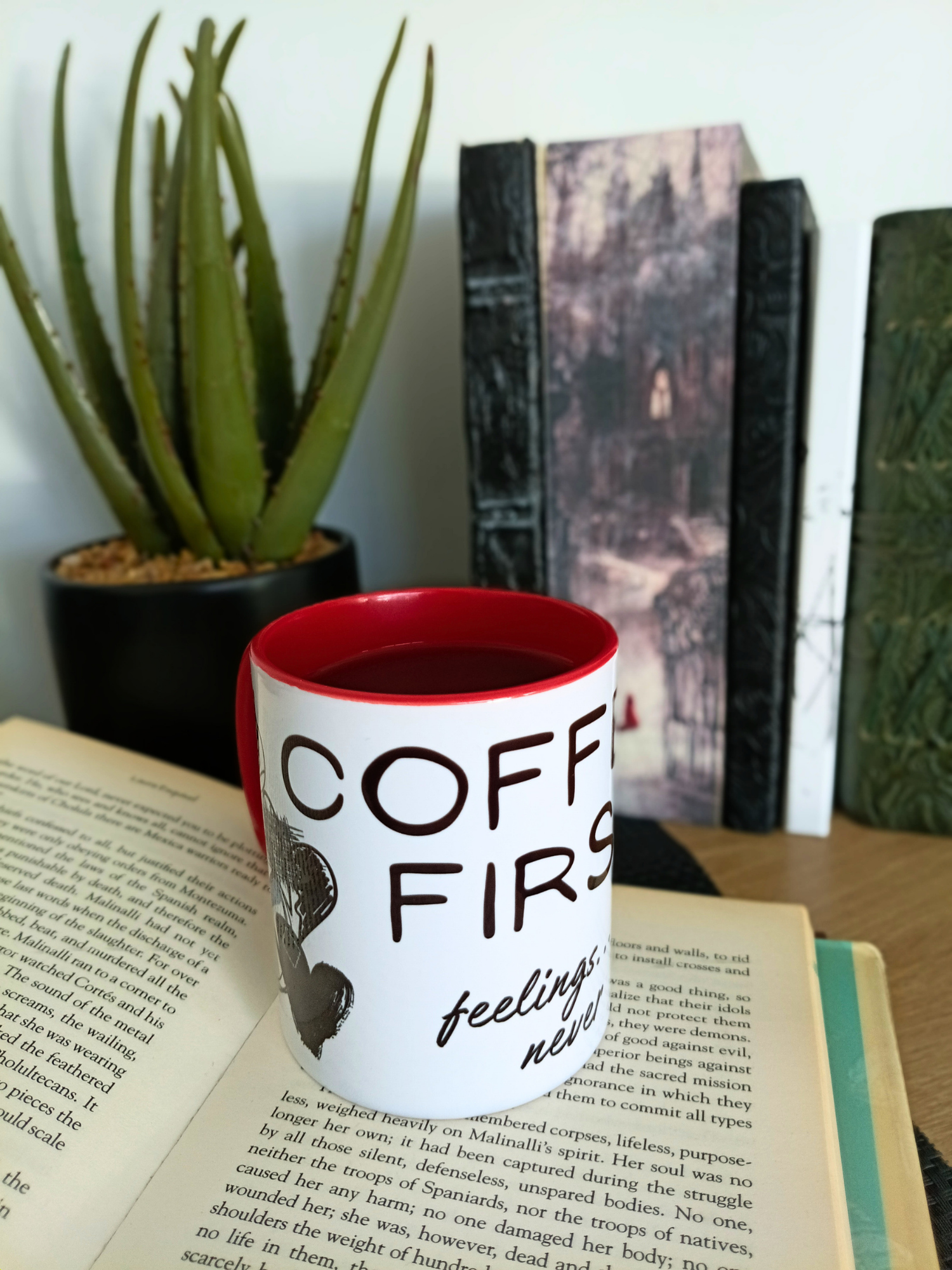 Mug with 'Coffee First, Feelings Later' text on a book with a plant and books in the background