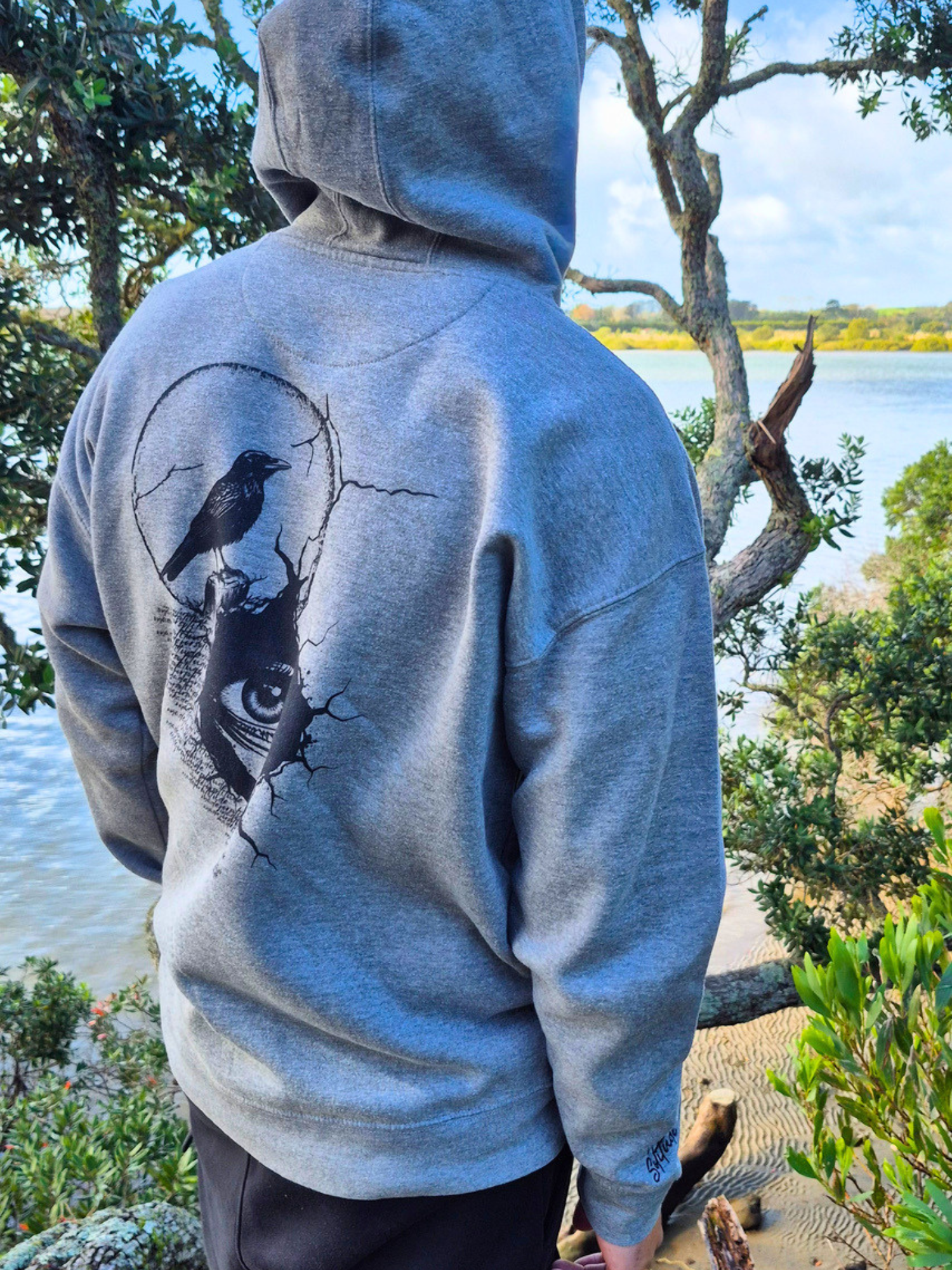 Person wearing a hoodie with a bird design outdoors by a body of water.