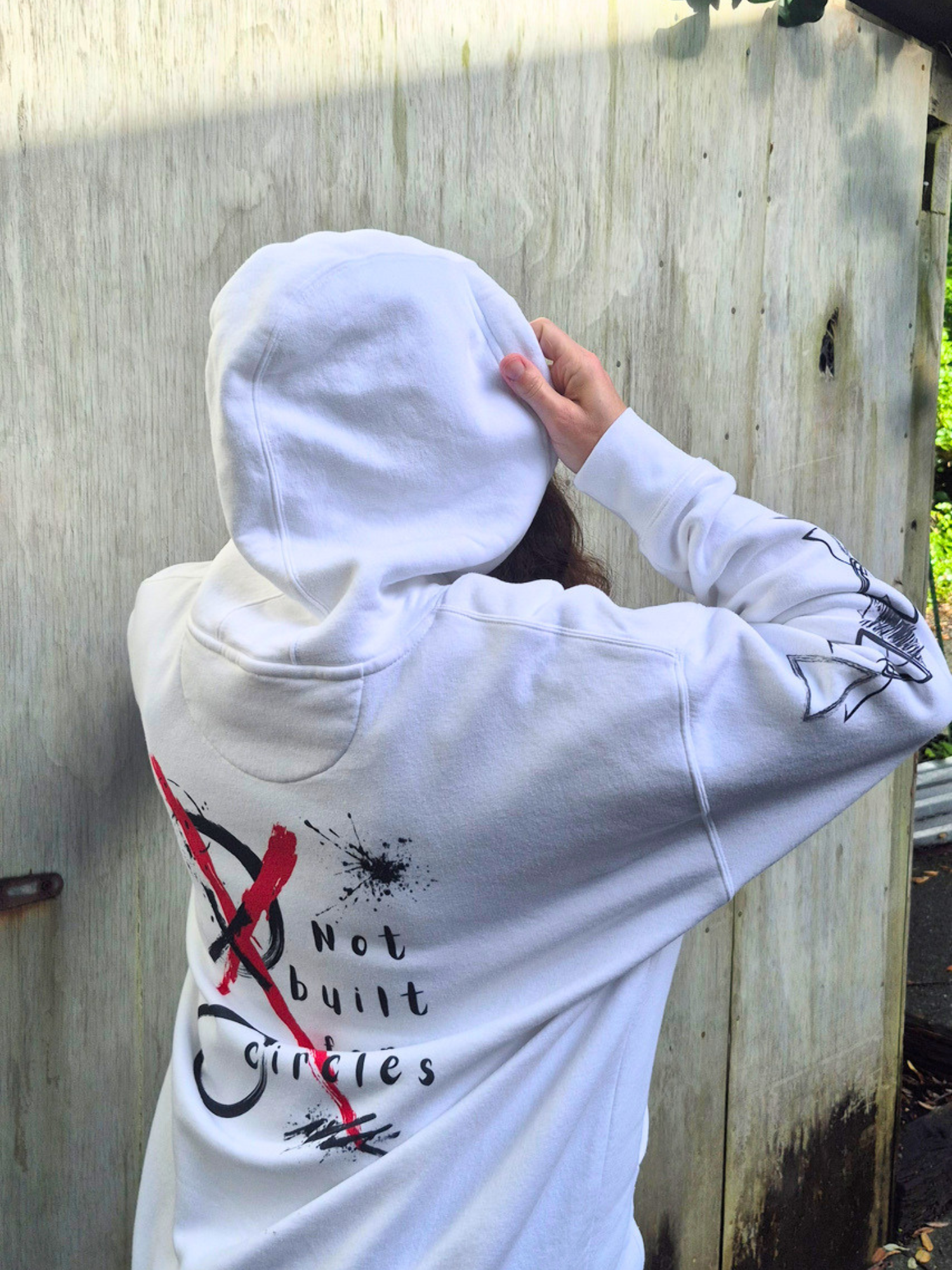 Person wearing a white hoodie with a graphic design and text, standing against a wooden fence.